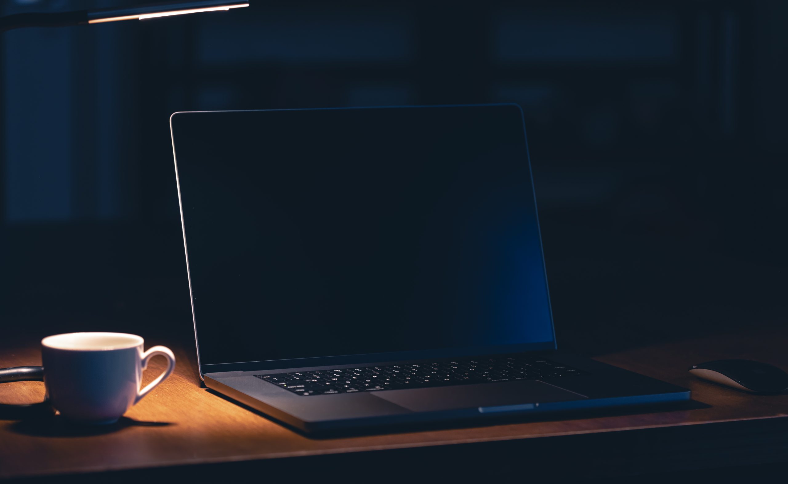 Klēpjdators uz galda blakus kafijas krūzei tumšā telpā ar galda lampu