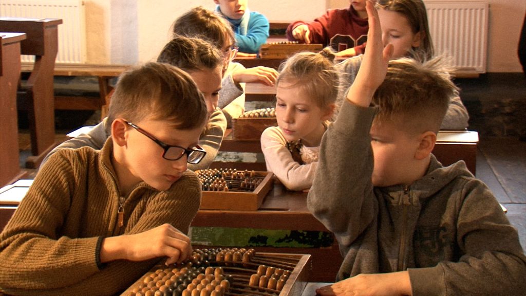 Grupa skolēnu sēž pie galdiem ar skaitāmmašīnām. Viens zēns paceļ roku.