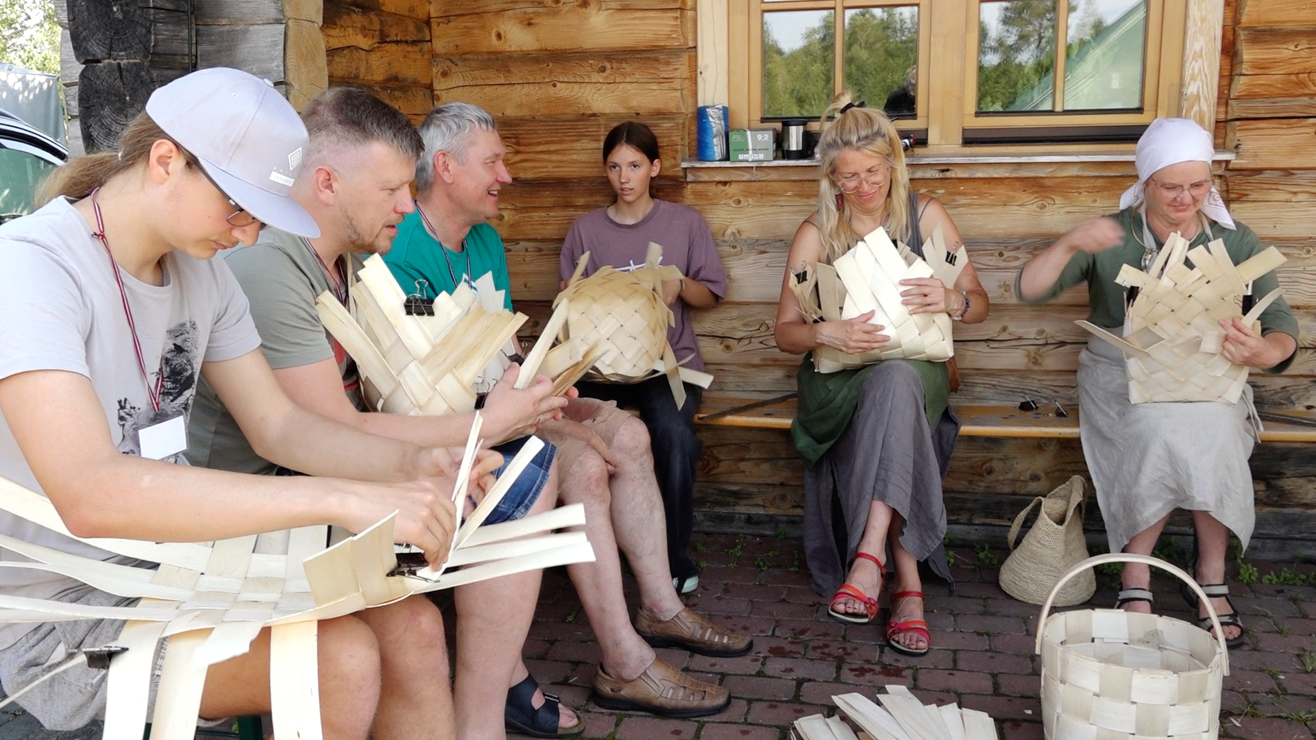 Dalībnieki saietā “Trejdeviņi” piedalās grozu pīšanas darbnīcā pie koka mājas.