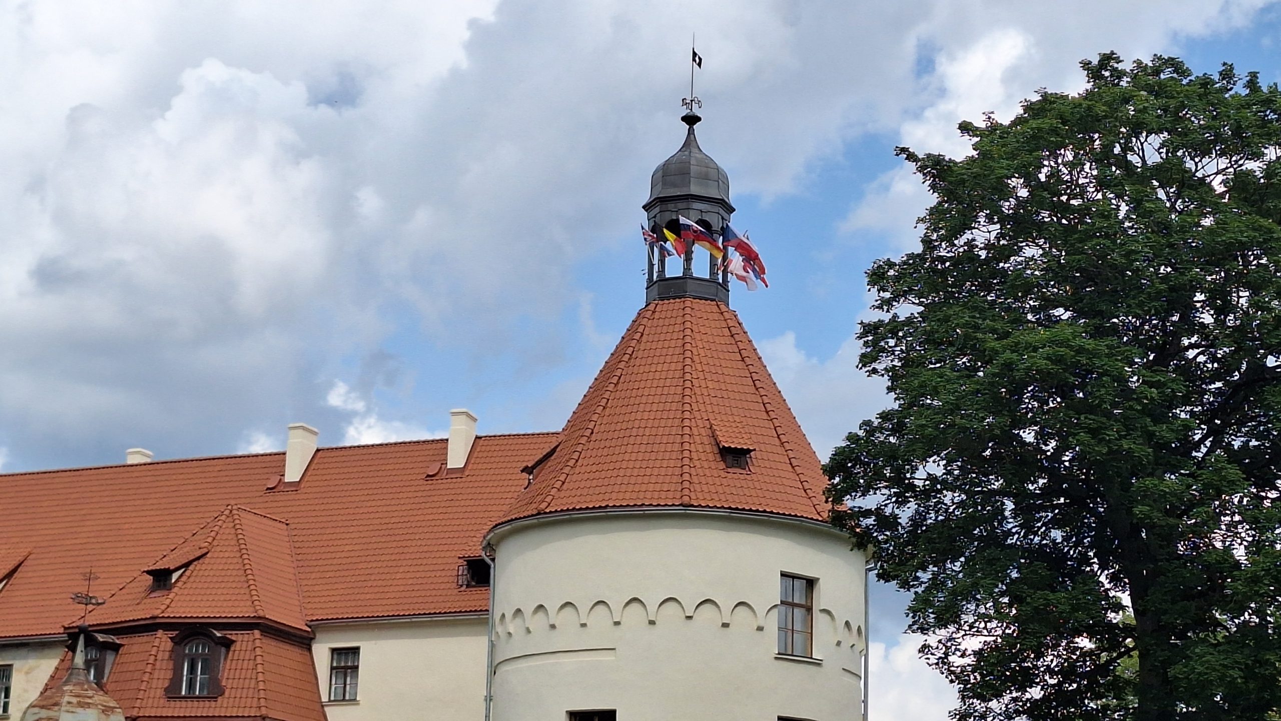 Jaunpils pils tornis ar plīvojošiem karogiem mākoņainās debesīs.