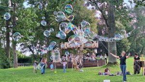 Bērni un pieaugušie parkā pie karuseļa spēlējas ar lieliem ziepju burbuļiem.