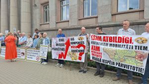 Protests Latvijā ar dalībniekiem, kuri tur plakātus par sabiedriskā transporta finansējumu un izmaksu pieaugumu.