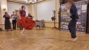 Vīrietis un sieviete dejo telpā ar koka grīdu, uz kuras ir krustveida marķējums.