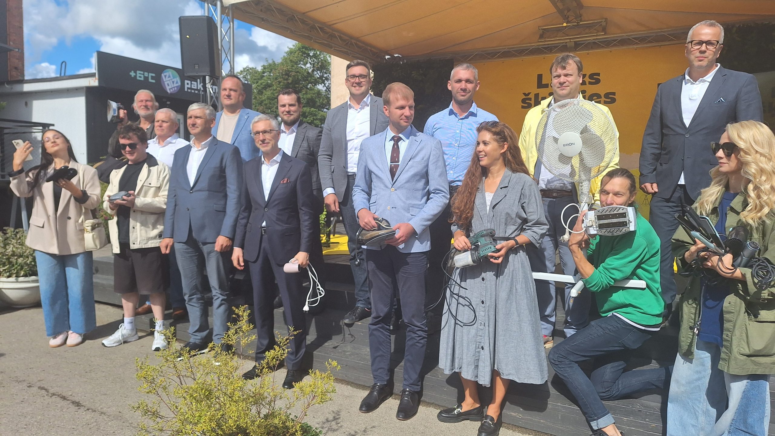 Cilvēku grupa, tostarp ministrs, pozē uz skatuves, turot vecas elektroierīces.