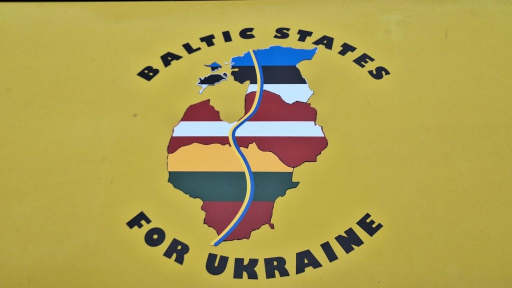 Uz dzeltenas virsmas ir attēlots stilizēts Baltijas valstu kartes siluets ar to karogu krāsām un divi uzraksti "BALTIC STATES" un "FOR UKRAINE".