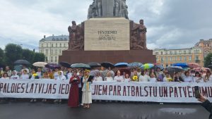 Cilvēku grupa ar lietussargiem stāv Brīvības pieminekļa priekšā, turot plakātu.