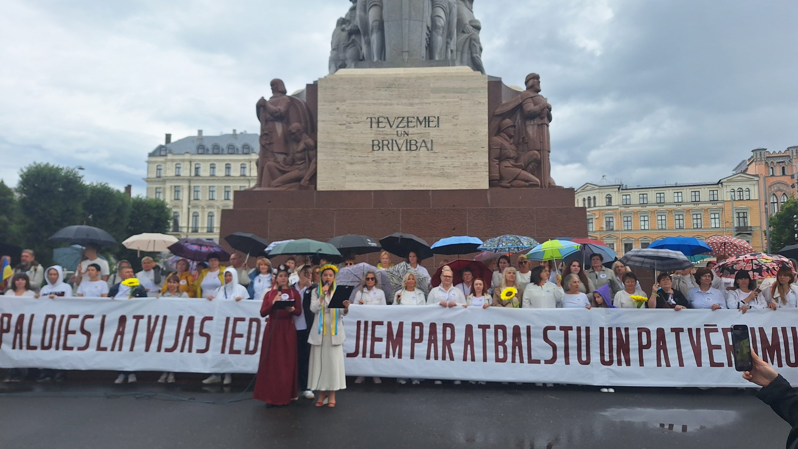 Cilvēku grupa ar lietussargiem stāv Brīvības pieminekļa priekšā, turot plakātu.