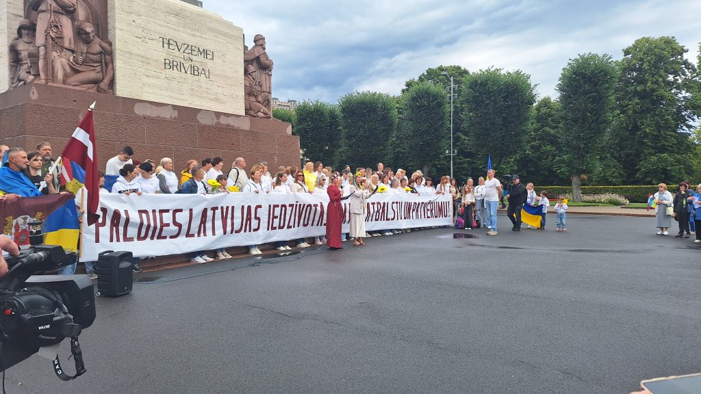 Liela cilvēku grupa stāv pie Brīvības pieminekļa, turot baltu plakātu.