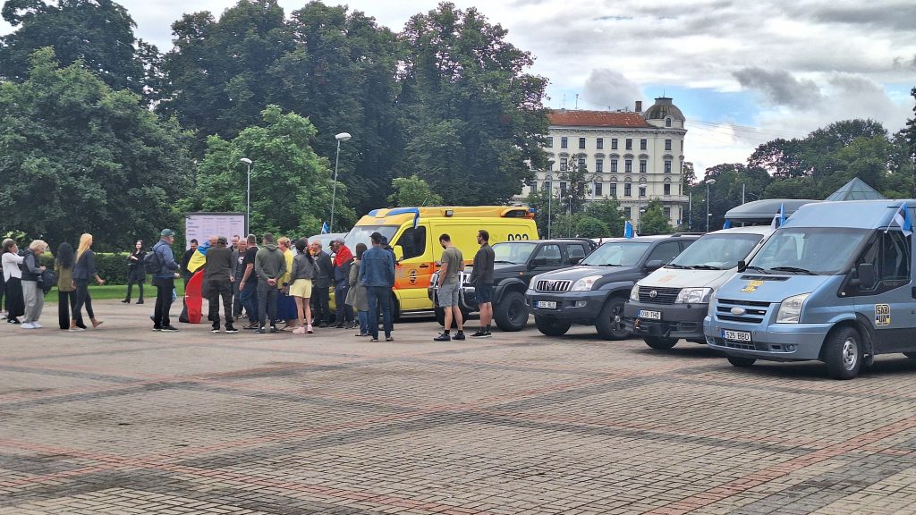 Cilvēku grupa pulcējas pie automašīnām, kas tiek sagatavotas nosūtīšanai uz Ukrainu.