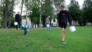Cilvēki, tostarp bērni, ar baltām spaiņu kastēm lasa Spānijas kailgliemežus zālājā pie kokiem.