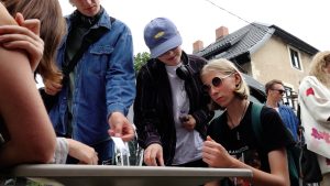 Jaunieši ārā pie galda piedalās aktivitātē, skatoties un pārrunājot uzdevuma kartītes.