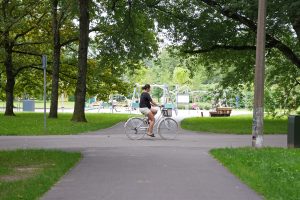Sieviete brauc ar velosipēdu pa asfaltētu celiņu Rīgas Uzvaras parkā.