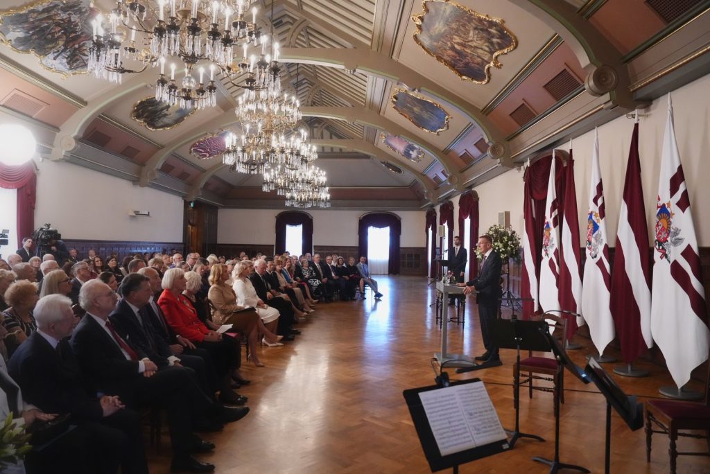 Vairāki cilvēki sēž rindās svinīgā zālē, klausoties runātāju.