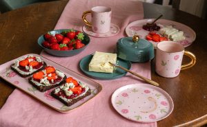 Katrīnas Keramikas porcelāna krūzes un šķīvji ar zelta rokturiem, zemenes un siermaizes uz galda