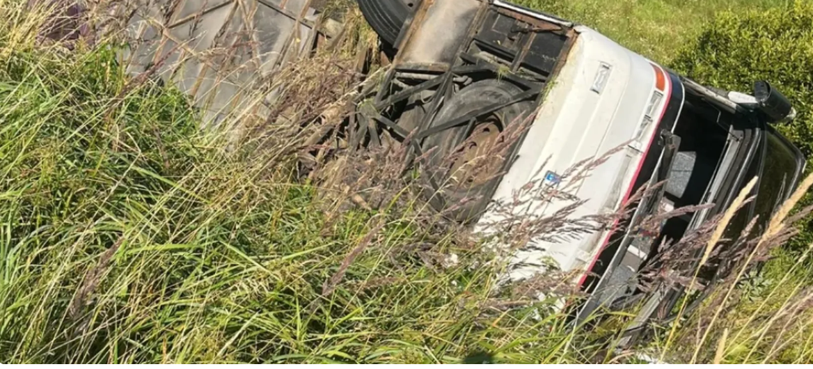 Avārijā cietis pasažieru autobuss guļ uz sāniem zālē pēc nobraukšanas no ceļa.
