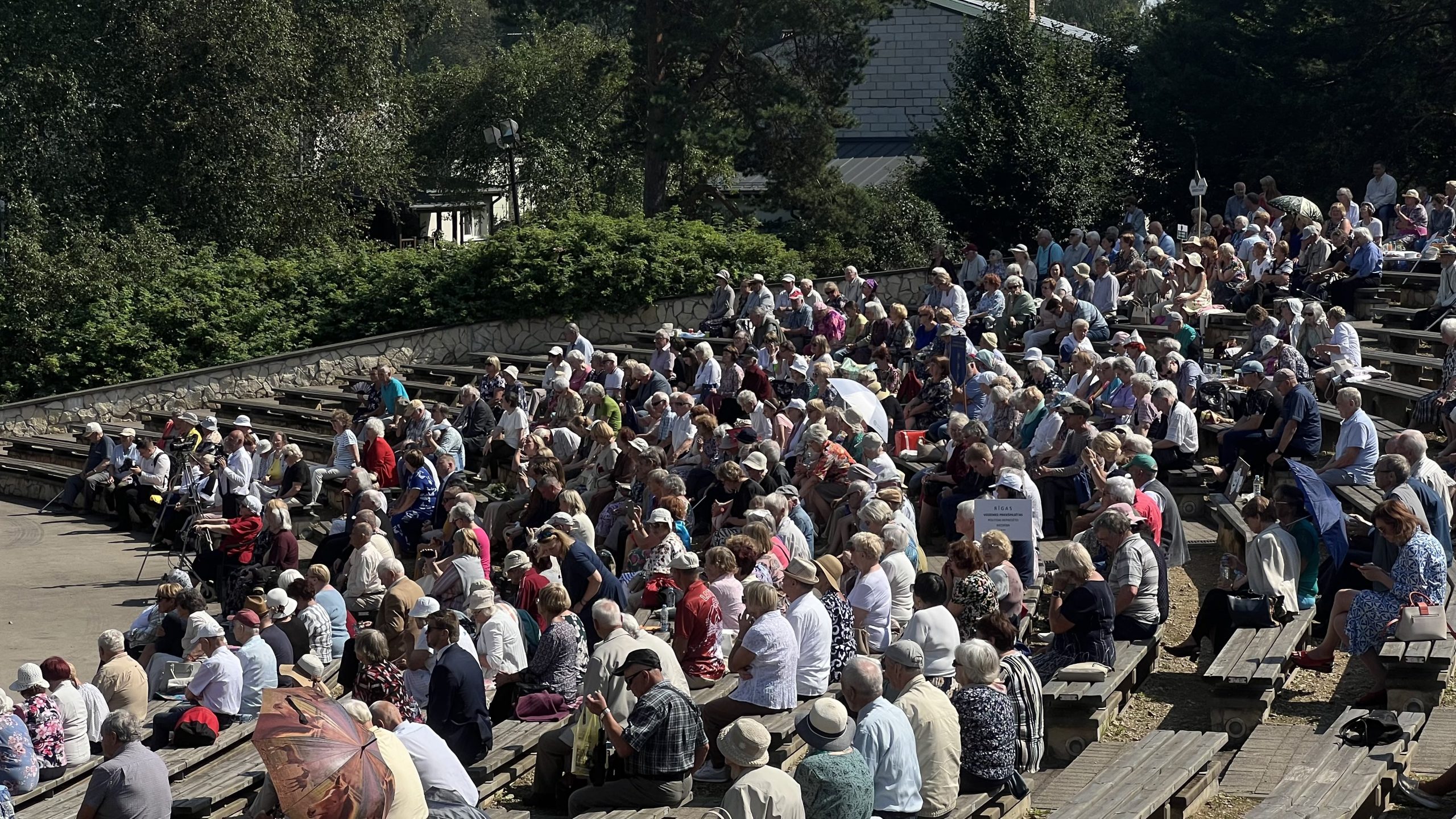 Cilvēku pūlis brīvdabas estrādē saulainā dienā.
