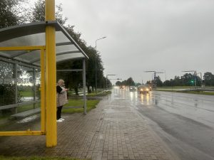 Sieviete stāv autobusa pieturā lietainā dienā pie ielas ar satiksmi