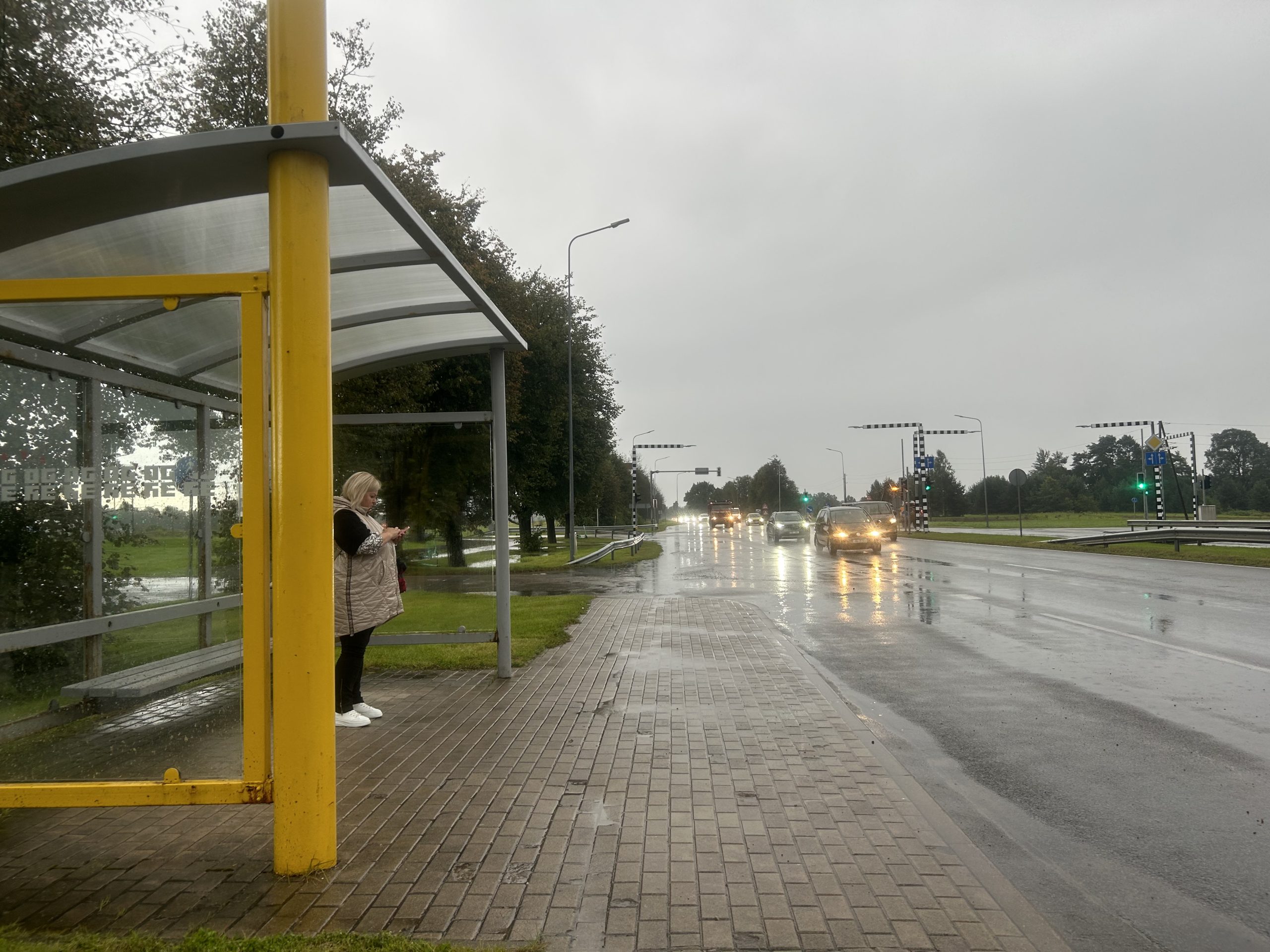 Sieviete stāv autobusa pieturā lietainā dienā pie ielas ar satiksmi