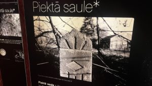 Betona stabs ar stilizētu saules staru un romba ornamentu pie žoga, rīta gaismā.