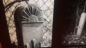 Vecs akmens stabs ar pusapaļu saules staru ornamentu pie žoga.