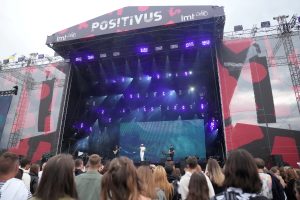 Mūzikas festivāla "Positivus" galvenā skatuve ar spilgtu apgaismojumu un priekšnesumu, ko vēro liels skatītāju pūlis.