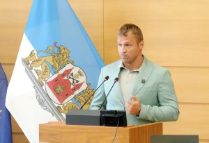 Mairis Briedis stāv pie mikrofoniem Rīgas domes zālē, runājot ar nopietnu sejas izteiksmi