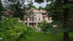 Lielplatones muižas ēka aiz kokiem un krūmiem vasaras zaļumā