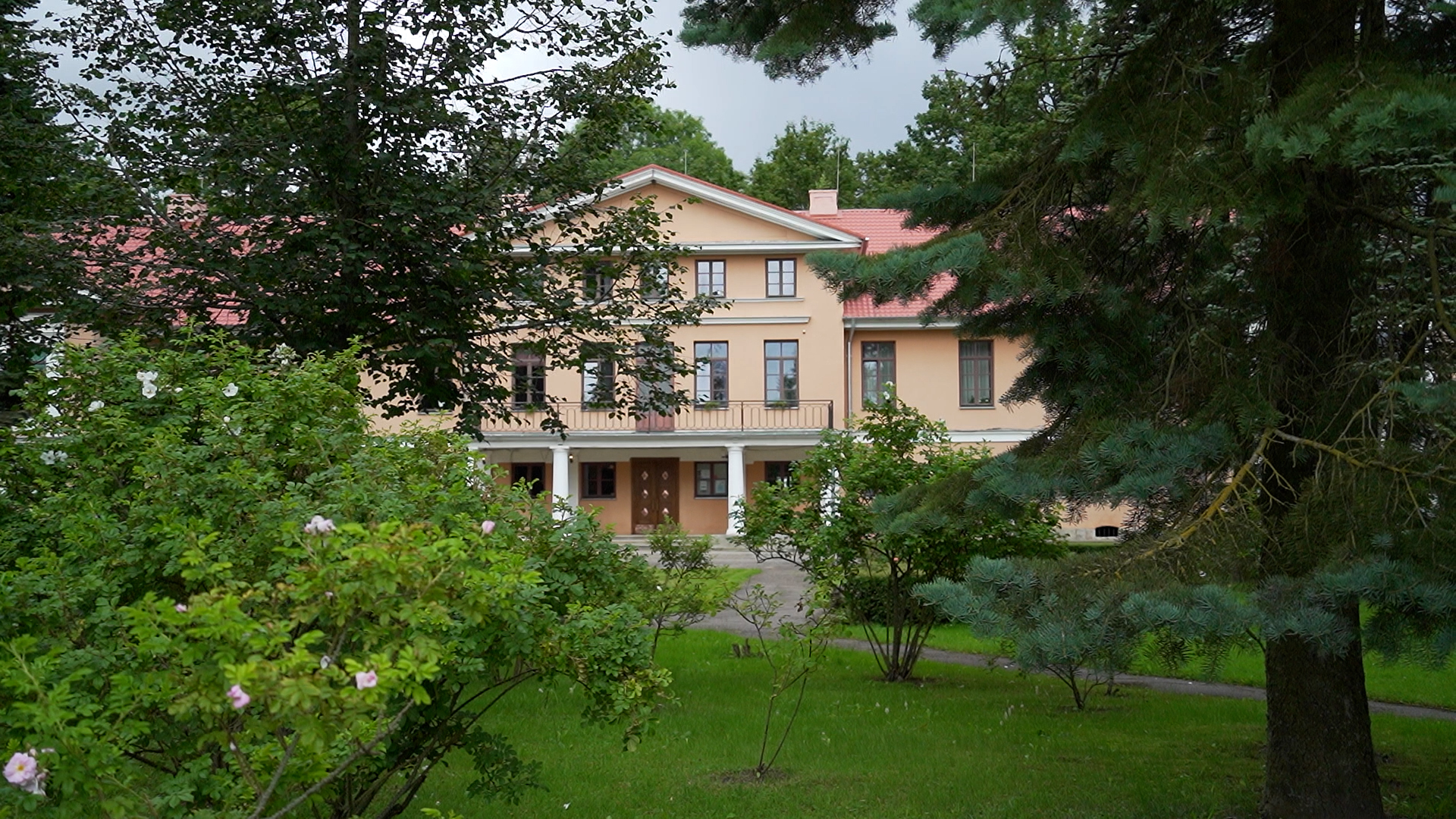 Lielplatones muižas ēka aiz kokiem un krūmiem vasaras zaļumā