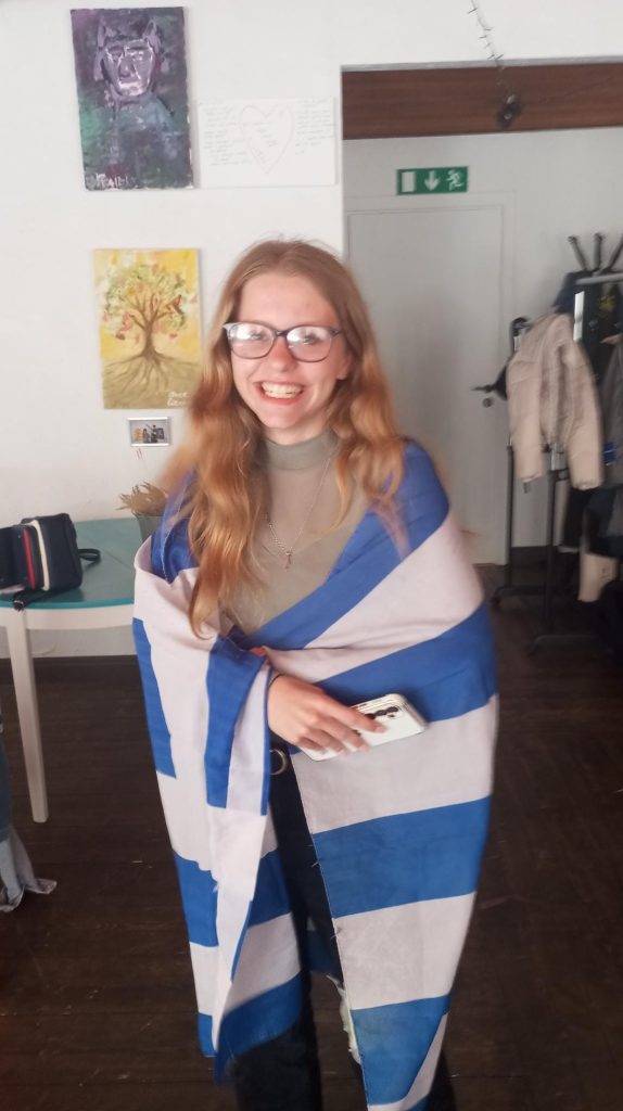 Jauniete smaidot stāv telpā, aptinusies ar karogu ar zilām un baltām svītrām
