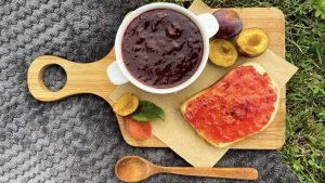 Plūmju ievārījums bļodiņā ar grauzdētu maizi un svaigām plūmēm uz dēlīša.