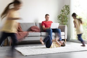 Tēvs mēģina strādāt ar portatīvo datoru, sēžot uz dīvāna, kamēr viņa bērni rotaļājas