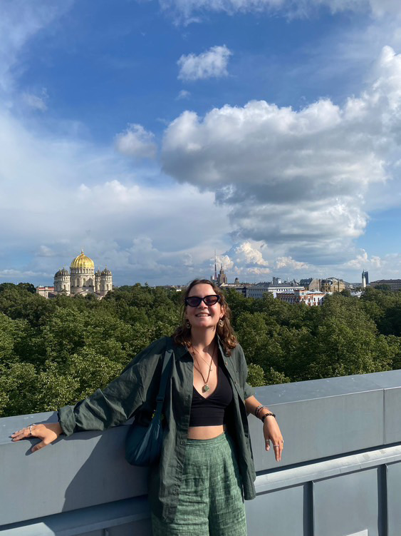 Mona Treija smaida, stāvot uz tilta ar Rīgas panorāmu fonā, kur redzama pareizticīgo katedrāle.