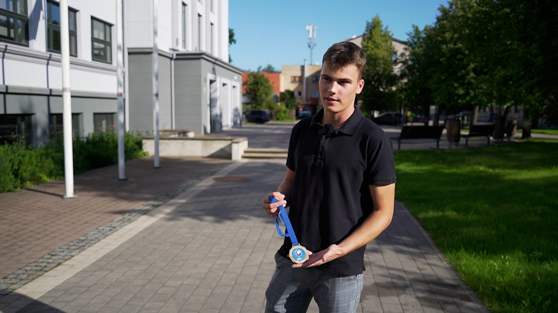 Jaunietis ar medaļu rokās stāv pilsētvidē pie ēkas.