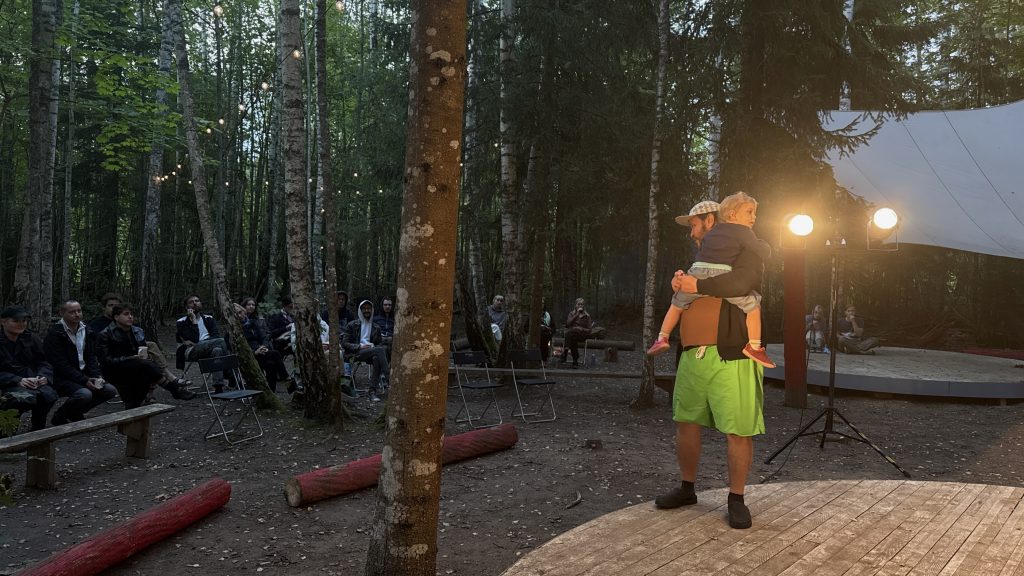 Vīrietis ar bērnu uz rokām stāv uz skatuves meža vidū, vērojot publiku