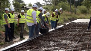 Cilvēki ar ķiverēm un atstarojošām vestēm betonē būves pamatus