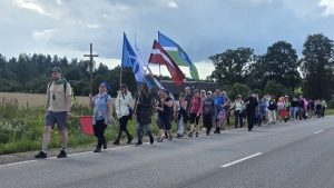 Svētceļnieku grupa ar karogiem soļo pa ceļu lauku apvidū