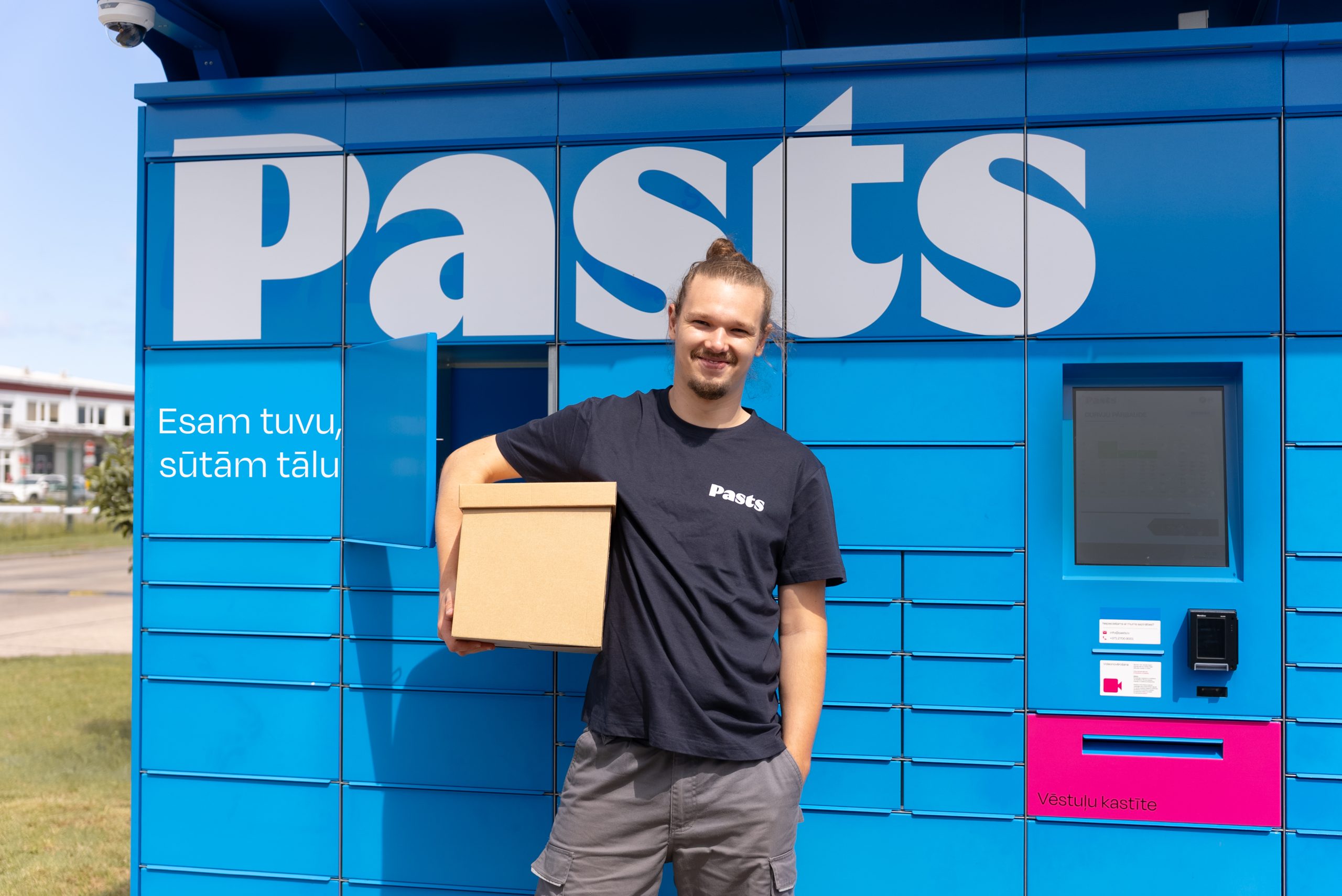 Jauns vīrietis smaidot stāv pie zila Latvijas Pasta pakomāta ar brūnu sūtījuma kasti rokās.