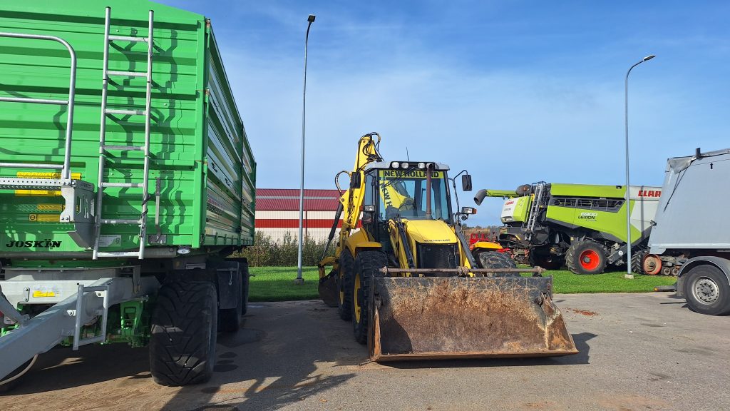 Dzeltenas krāsas universālais iekrāvējs (New Holland) un zaļas krāsas graudu kombains (Claas) stāv blakus zaļai piekabes (Joskin) daļai uz asfaltēta laukuma.