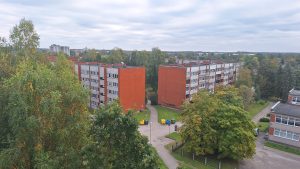 Daudzdzīvokļu dzīvojamo māju skats no augšas ar sarkaniem ķieģeļu elementiem, kas ieskauj zaļi koki.