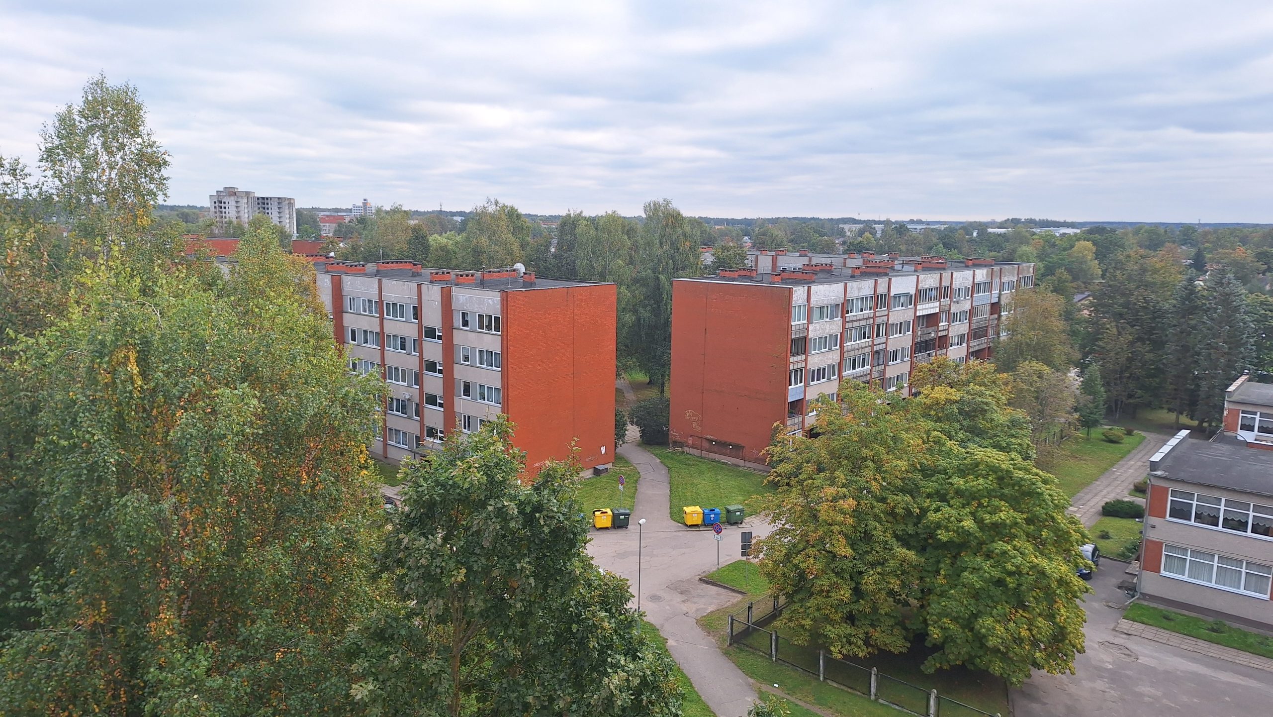 Daudzdzīvokļu dzīvojamo māju skats no augšas ar sarkaniem ķieģeļu elementiem, kas ieskauj zaļi koki.