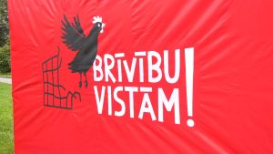 Sarkans plakāts ar uzrakstu “Brīvību vistām!” un vistas attēlu.