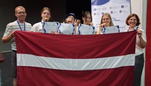 Četri skolēni un divi skolotāji pozē ar lielo Latvijas karogu Starptautiskajā angļu valodas olimpiādē Rumānijā.