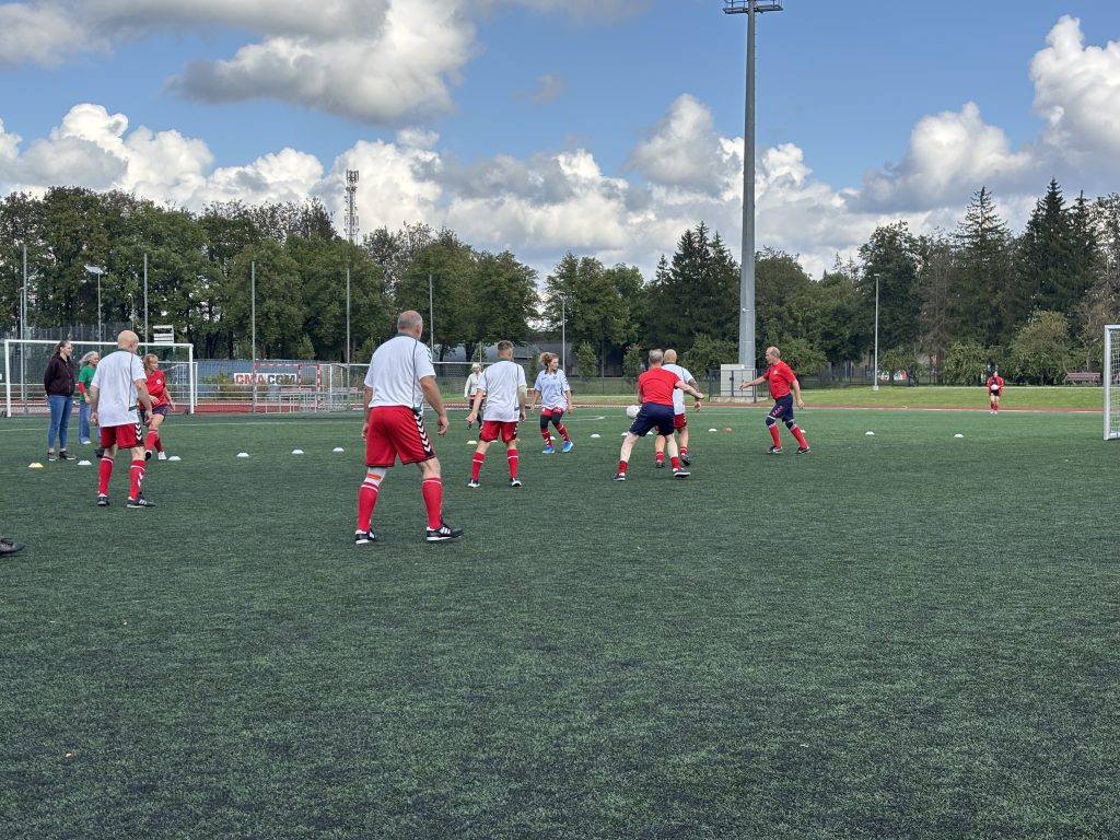 Soļbola spēlētāji trenējas uz futbola laukuma ar muguru pret kameru.