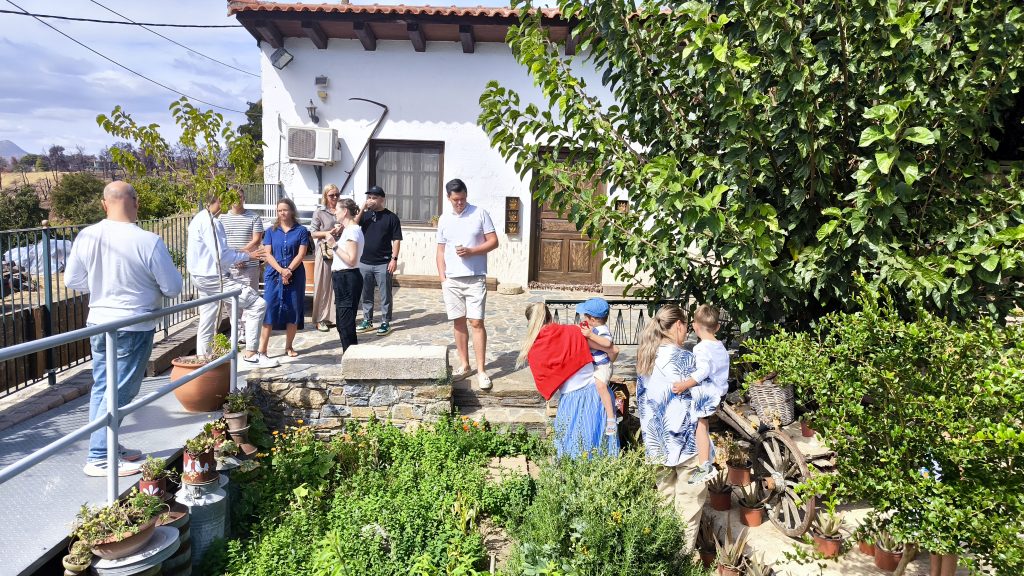 Latviešu ģimenes un bērni stāv pie Grieķijas Eiropas maizes muzeja ēkas, kas ir balta, lauku stila māja ar koka elementiem un bagātīgiem zaļumiem.