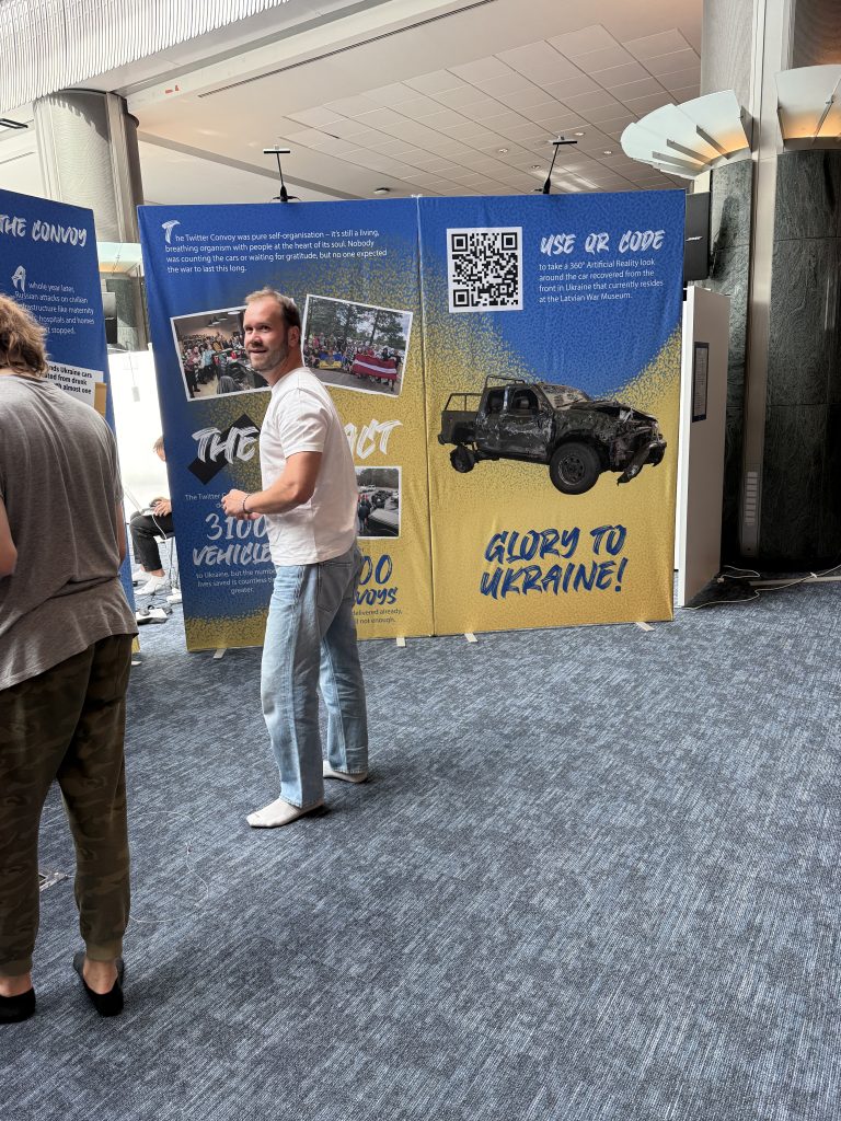 Vīrietis stāv pie ekspozīcijas stenda ar sadegušu automašīnu un QR kodu
