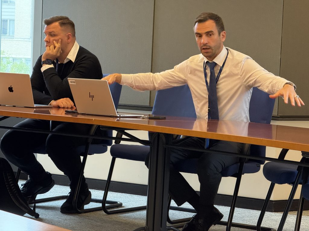 Divi vīrieši konferenču zālē. Viens no tiem ar žestiem stāsta.