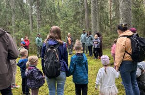 Cilvēku grupa stāv aplī meža klajumā, klausoties pavadoni. Priekšplānā bērni un pieaugušie ar mugursomām.