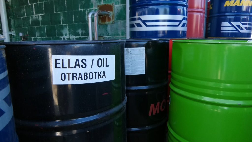 Liela melna muca ar uzrakstu "ELLAS / OIL OTRABOTKA" un citas krāsainas mucas fonā.