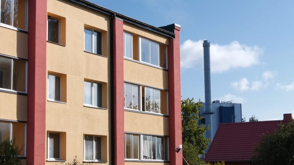 Daudzdzīvokļu ēkas dzeltenā fasāde ar sarkanām apmalēm; fonā redzams siltumcentrāles skurstenis.