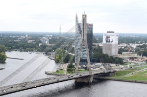 Panorāmas skats uz Vanšu tiltu Rīgā, šķērsojot Daugavu, ar pilsētas siluetu fonā un intensīvu satiksmi uz tilta.